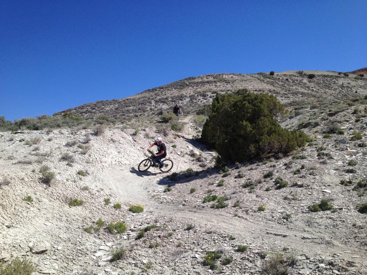 A person riding a mountain bike down a winding trail on a sandy hillside, surrounded by sparse vegetation and shrubs, under a clear blue sky. Another cyclist can be seen in the background, navigating the terrain. Tnt mountain bike trail.
