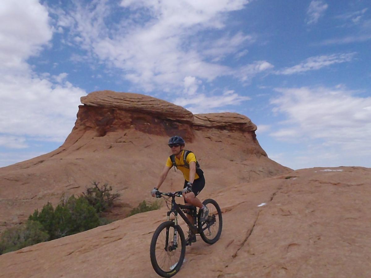 Jamis Dakar XLT: A person riding a mountain bike on rocky terrain with a unique rock formation in the background, under a partly cloudy sky.