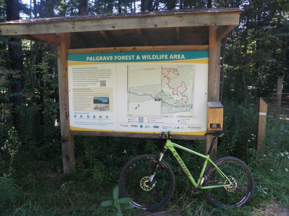 A mountain bike is parked next to a wooden sign for the Palgrave Forest & Wildlife Area, which features a detailed map of the trails and information about the area. The sign is surrounded by dense greenery and wildflowers, indicating a natural setting for outdoor activities. Palgrave Trail mountain bike trail.