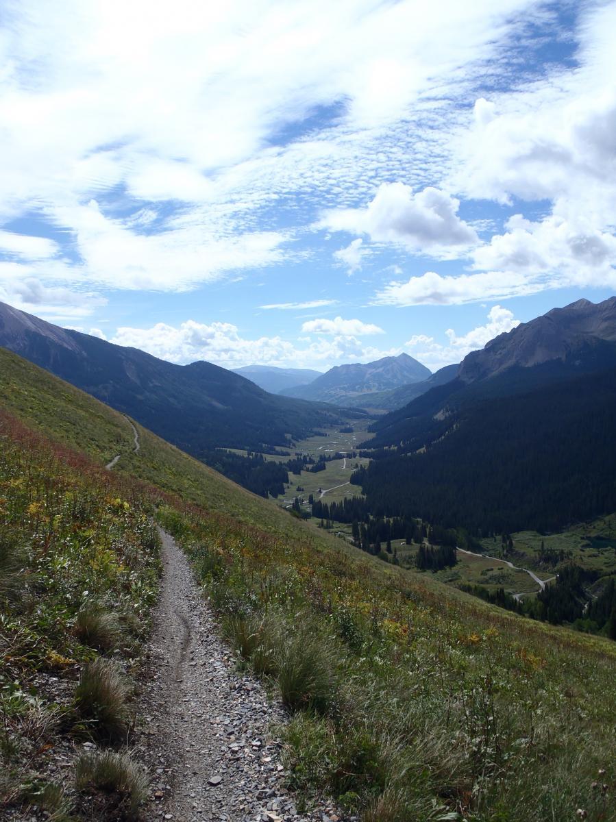 A winding dirt path leads downhill through grassy terrain, surrounded by mountains under a partly cloudy sky. The valley below features lush forests and open fields with some roads visible in the distance. Trail 401 mountain bike trail.