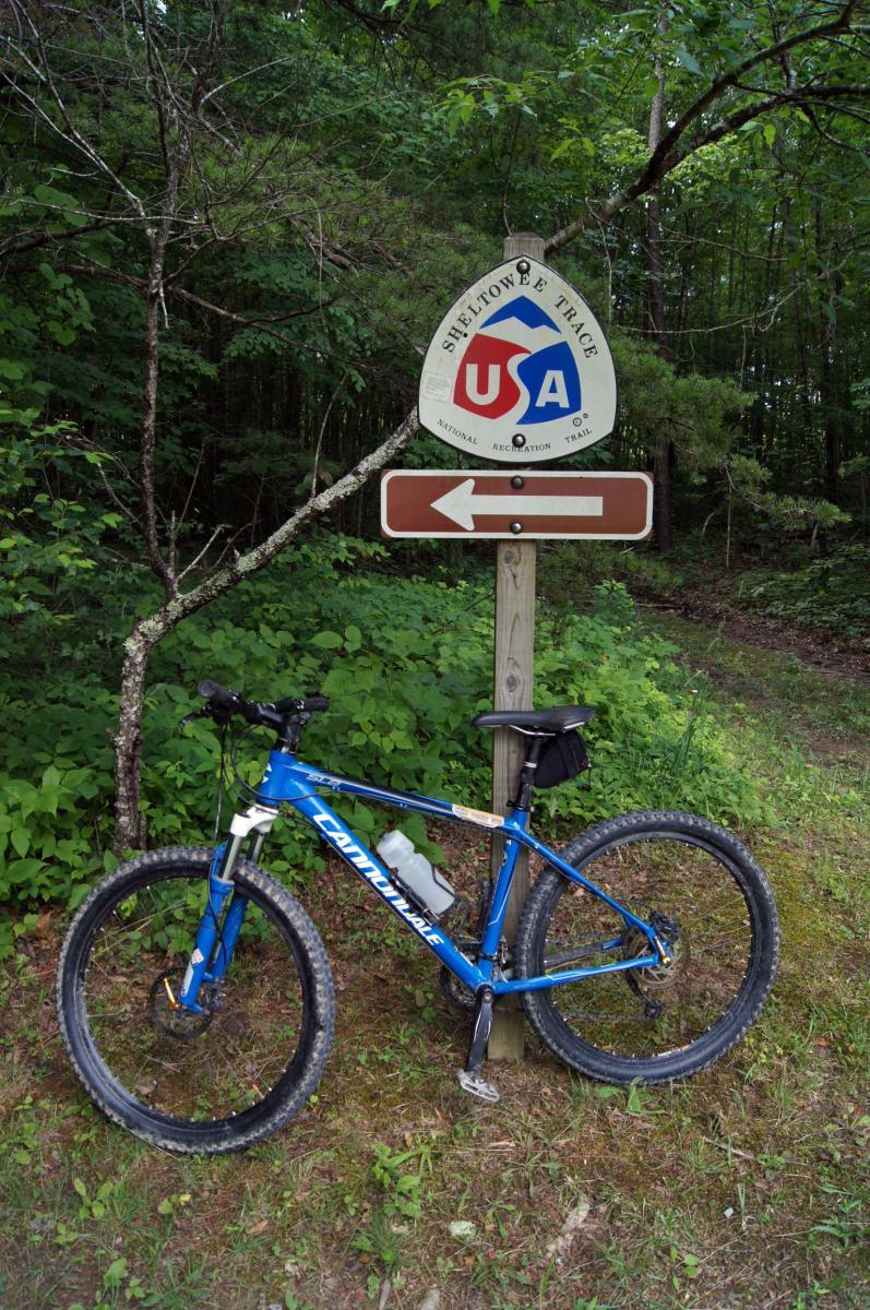 A blue mountain bike is parked beside a sign for the Sheltowee Trace National Recreation Trail, indicating a left turn. The background features lush greenery and trees typical of a wooded area. Sheltowee Trace mountain bike trail.