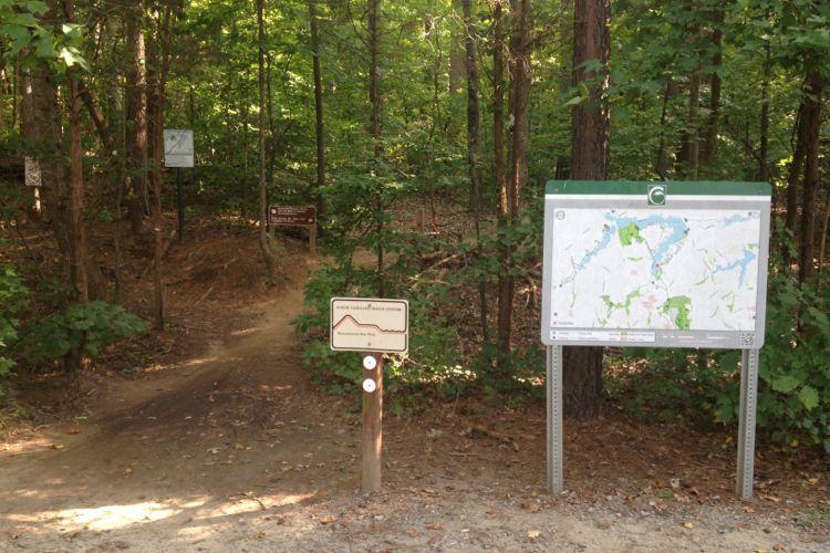 A wooded trail intersection with a dirt path leading into the forest. A trail map sign stands at the right, while a wooden post with trail information is on the left. Various informational signs are partially visible in the background among the trees. The scene is surrounded by green foliage, indicating a sunny day. Wild Turkey mountain bike trail.