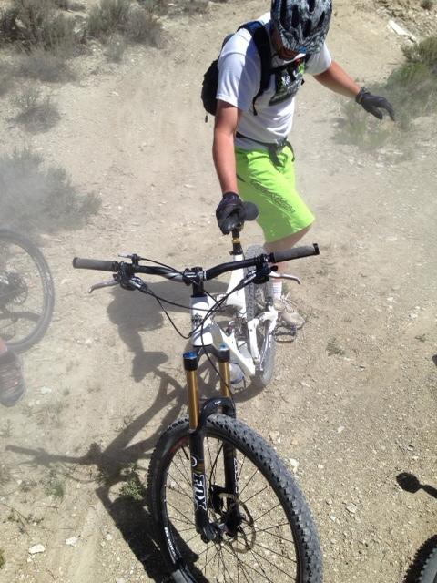 A person in a white shirt and green shorts adjusts the front tire of a white mountain bike on a dusty trail surrounded by sparse vegetation. The scene suggests an outdoor biking activity in a rugged environment. Tnt mountain bike trail.