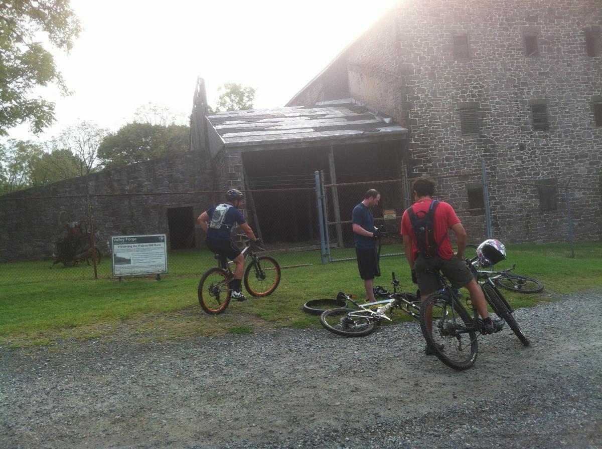 Three mountain bikers are gathered outside an old stone building at Valley Forge. One rider is seated on a bike, while another is checking equipment, and the third is standing nearby. A sign in front of the building provides information about the historic site. The scene is set in a grassy area with gravel paths and trees in the background. Valley Forge Park River Trail mountain bike trail.