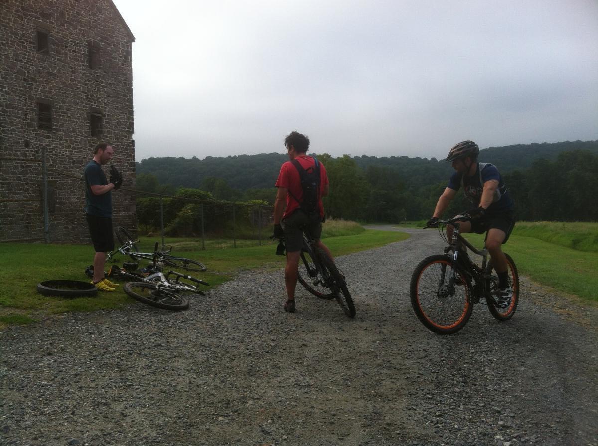 Three mountain bikers in a rural setting near a stone building are adjusting their bikes and gear. Two riders are standing on gravel, one wearing a red shirt and the other in a blue shirt with a helmet. A third rider is seen in motion on a bike. The background features green hills under a cloudy sky. Valley Forge Park River Trail mountain bike trail.