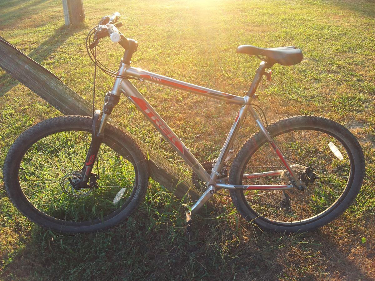 Trek 3500: A silver and red mountain bike resting on grass near a wooden fence, with the sun setting in the background, casting a warm glow over the scene.
