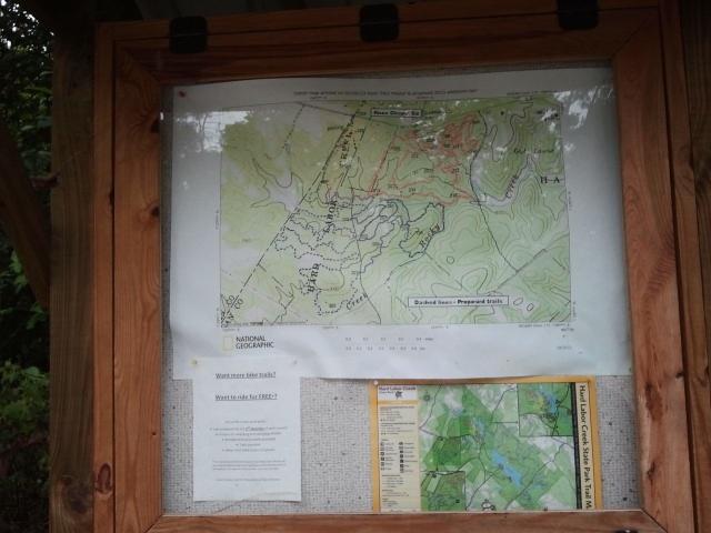 A wooden information board displaying a topographic map of a natural area, produced by National Geographic. The map outlines trails and geographical features, accompanied by informational text about hiking options and park guidelines. A smaller map is also visible on the bottom right of the board. Hard Labor Creek State Park mountain bike trail.