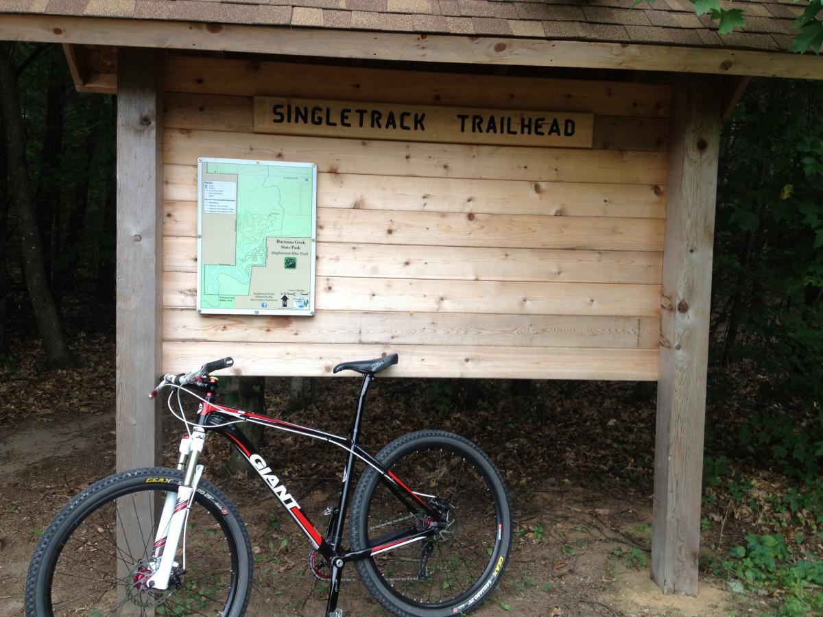 Giant Talon 29er: A mountain bike leaning against a wooden trailhead sign labeled "Singletrack Trailhead," featuring a map of the trail area. The surroundings include trees and earthy ground, indicating a natural outdoor setting.