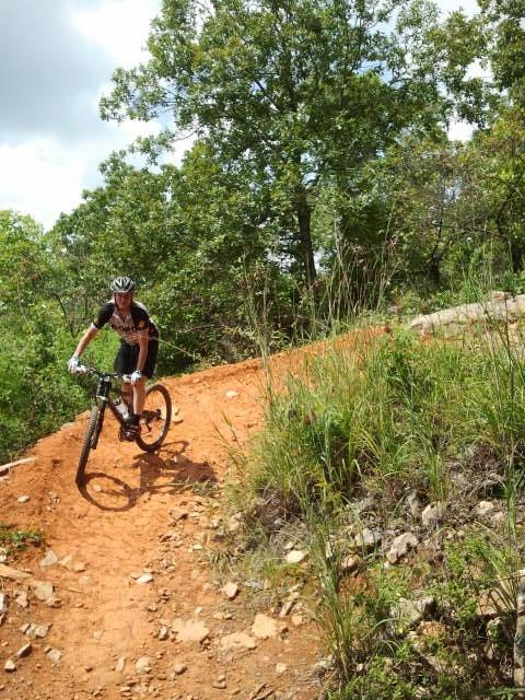 A mountain biker navigating a winding dirt trail surrounded by greenery and trees on a sunny day. Coldwater Mountain mountain bike trail.