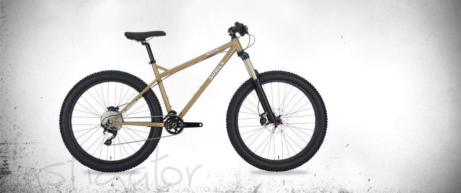 Surly Instigator: A mountain bike with a lightweight, tan-colored frame, equipped with wide tires, a front suspension fork, and black handlebars, set against a textured white background.
