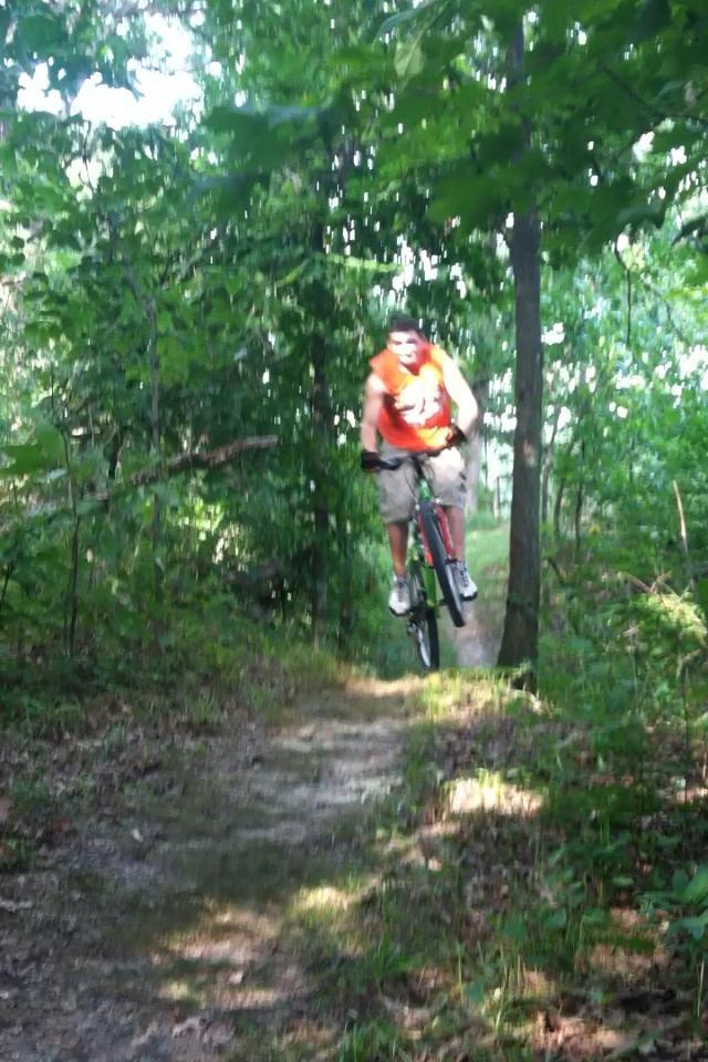 A person in an orange shirt jumps over a small dirt ramp while riding a mountain bike on a wooded trail, surrounded by green foliage. Camp Camfield mountain bike trail.