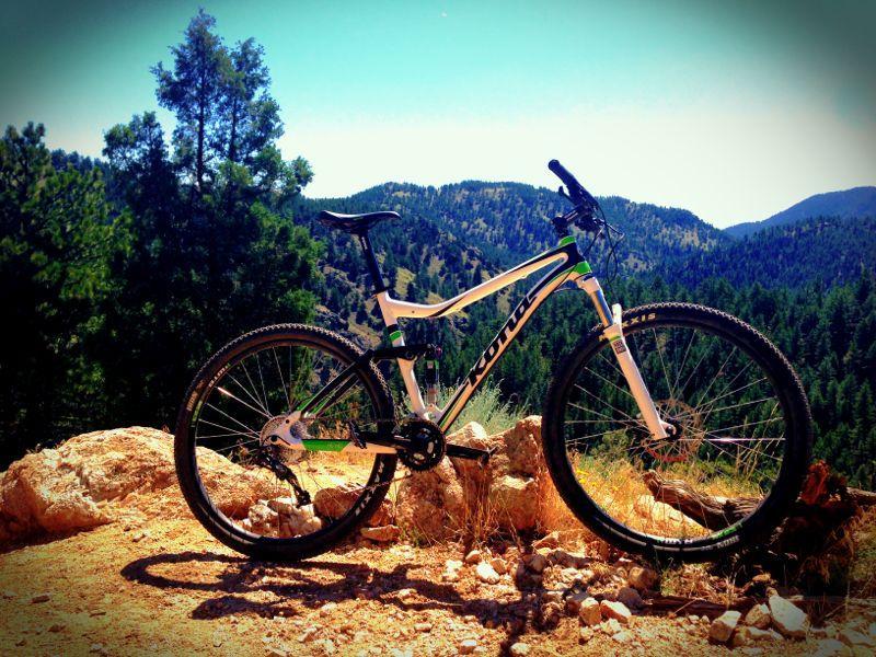 A mountain bike resting on rocky terrain, surrounded by lush green trees and rolling hills under a clear blue sky. The bike features a sleek white and black design with green accents, showcasing its robust tires and gear system, set against a backdrop of scenic mountainous landscape. Betasso Preserve mountain bike trail.