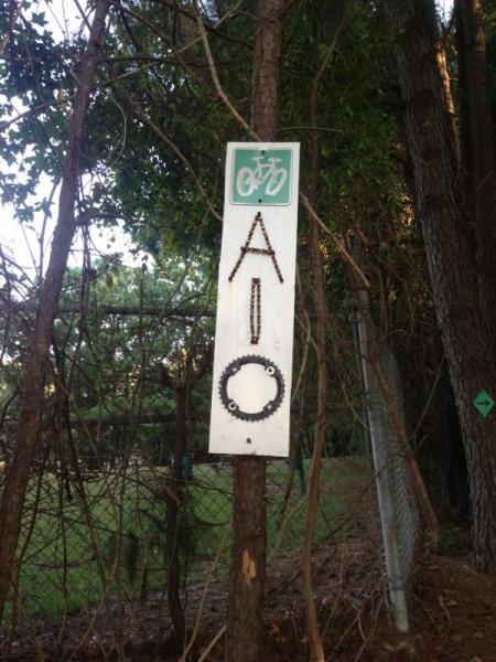 A sign mounted on a tree, featuring a bicycle symbol at the top and the letters "A I O" created with small objects below. The sign is surrounded by trees and a fence, indicating a wooded area. A-10 mountain bike trail.