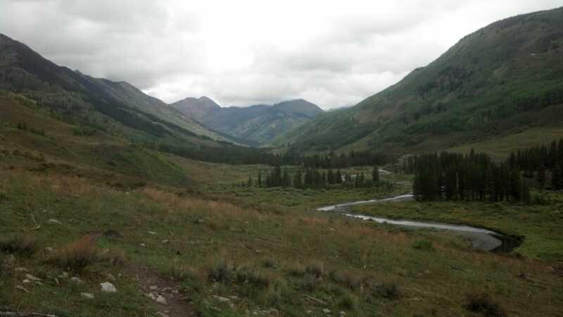 A scenic mountain landscape featuring rolling hills, lush green valleys, and a winding river under a cloudy sky. The foreground includes grassy areas with patches of trees, while the background showcases towering mountains with varied elevations. Lower Loop mountain bike trail.