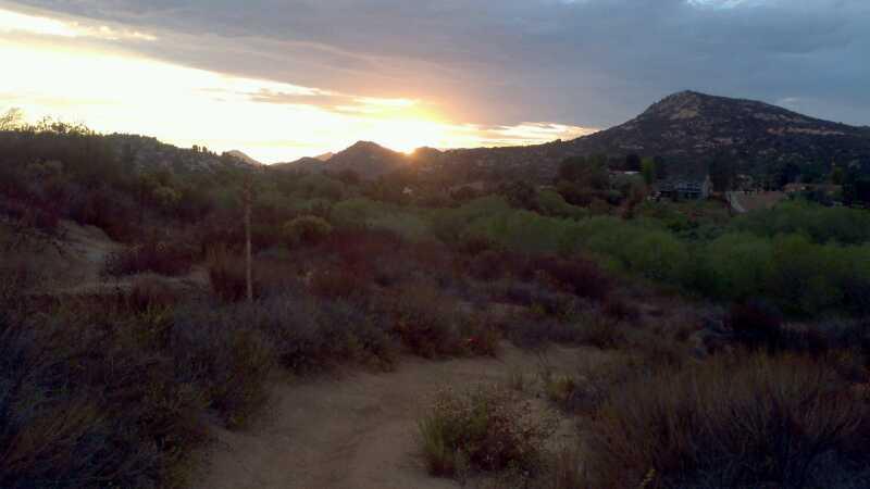 A scenic view of a mountainous landscape at sunset, showcasing a range of hills in the background under a cloudy sky. The foreground features a dirt path winding through patches of vegetation, including shrubs and low plants. The sun is setting behind the hills, casting warm hues across the sky and illuminating the scene with a soft glow. Happy