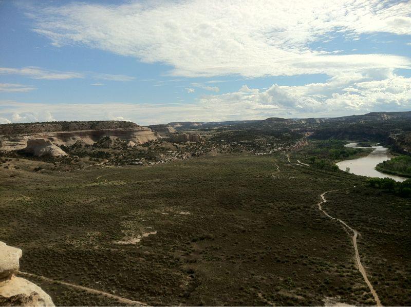 A scenic view of a wide, open landscape with rolling hills and a winding river. The sky is partly cloudy with blue patches peeking through. The foreground features green vegetation and a dirt path, while the background showcases rock formations and additional hills in the distance. Western Rim mountain bike trail.