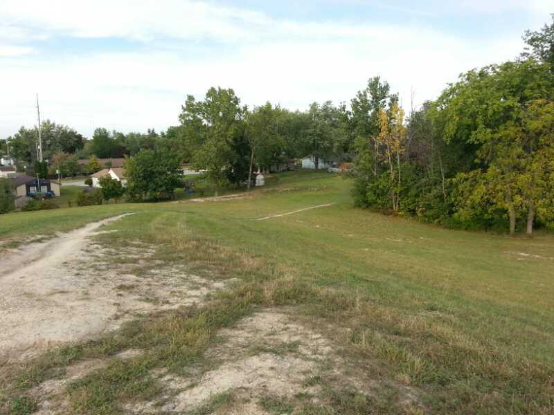 A view of a grassy slope leading down to a small neighborhood, surrounded by trees and houses. In the background, a dirt path winds through the grassy area, with a few structures visible among the greenery under a partly cloudy sky. Franke Park mountain bike trail.
