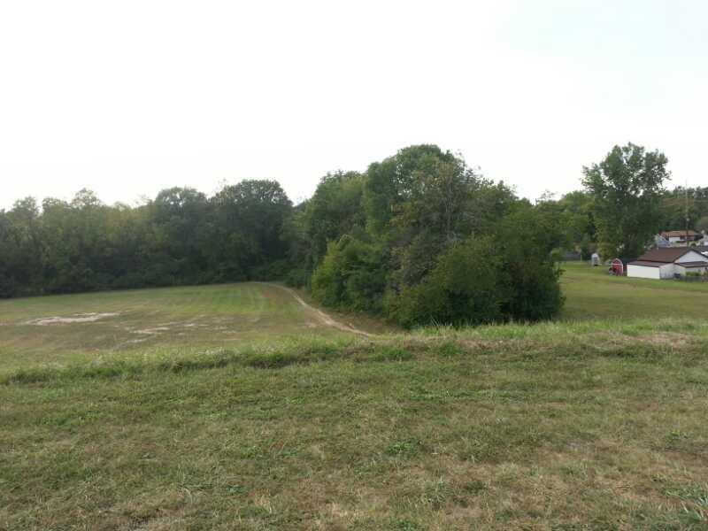A grassy area leads to a wooded edge with tall trees. In the distance, a few buildings and a red barn are visible, set against a cloudy sky. The landscape appears serene and rural. Franke Park mountain bike trail.