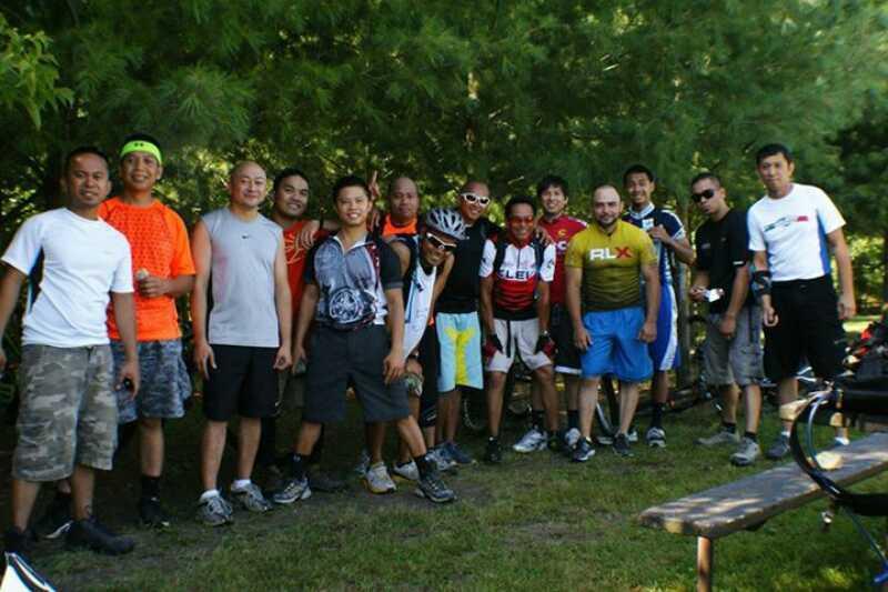 A diverse group of individuals posing together in a wooded area, dressed in athletic clothing, likely after a biking event. They are smiling and standing in a semi-circle, with bicycles visible in the background. The scenery features green trees and a picnic table. Kettle Moraine John Muir + Emma Carlin mountain bike trail.
