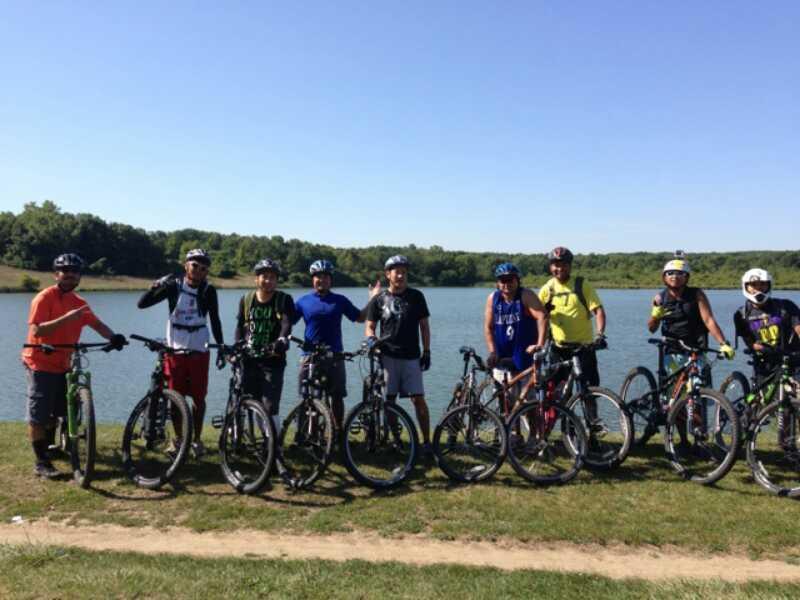 Group of eight cyclists in helmets posing with their mountain bikes by a lake on a sunny day, surrounded by greenery. Palos Forest Preserve mountain bike trail.