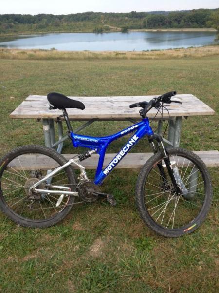 A blue Motobecane mountain bike is parked next to a wooden picnic table, set on a grassy area with a calm lake in the background. The bike has a silver frame with black wheels and is positioned to the right of the table, with trees and hills visible in the distance. Palos Forest Preserve mountain bike trail.
