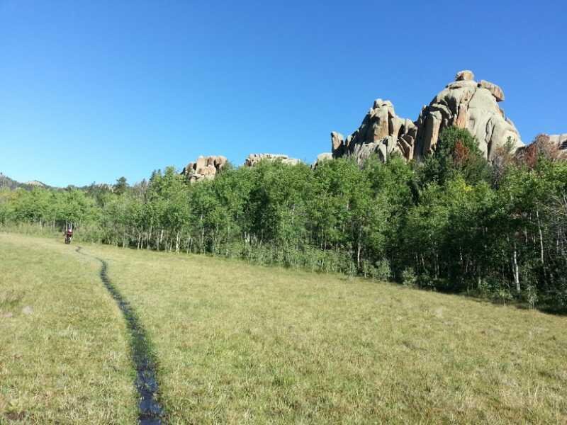 A scenic view of a grassy field with a winding dirt path leading through a grove of trees, set against a backdrop of large rock formations under a clear blue sky. Devil