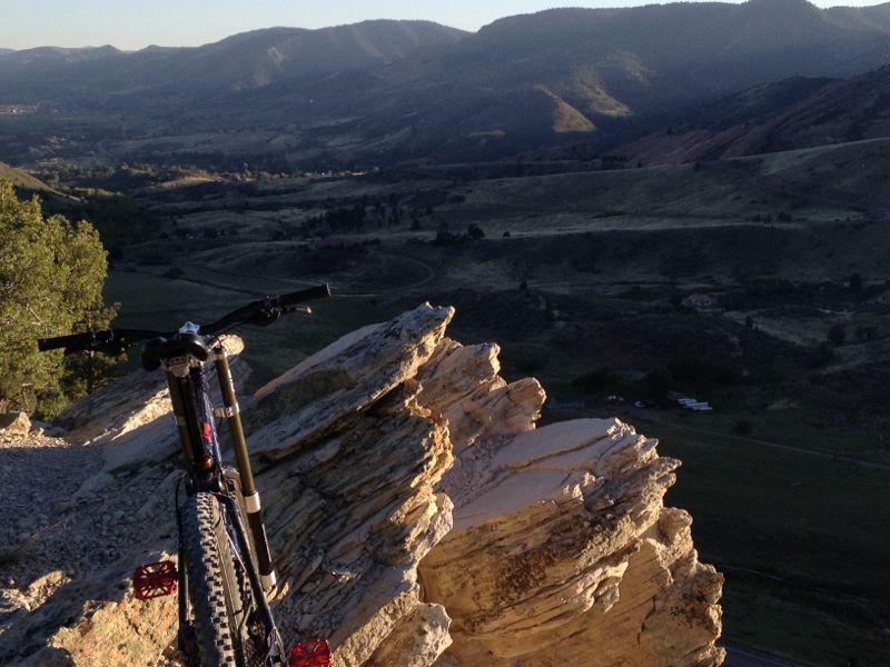 A mountain bike resting on a rocky outcrop, overlooking a scenic valley with rolling hills and distant mountains in the background, during sunset. Red Rocks / Dakota Ridge mountain bike trail.