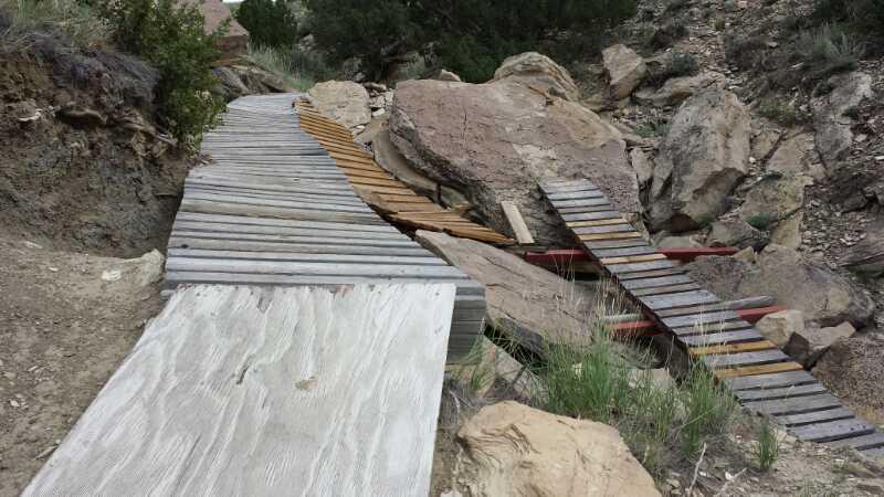 A broken and uneven wooden boardwalk crossing over rocky terrain, with large boulders and sparse vegetation visible on either side. The path appears to be damaged, with sections of the boardwalk missing or misaligned. South Shore Lake Pueblo mountain bike trail.