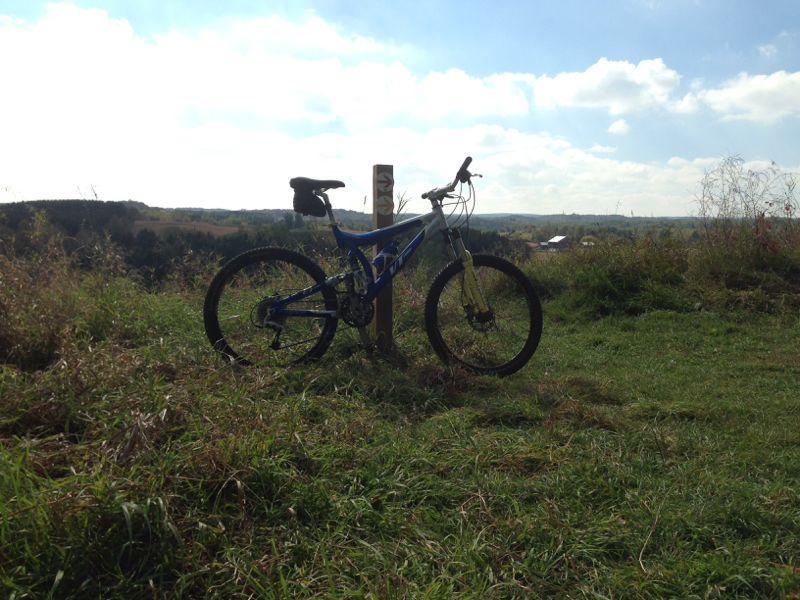 A blue mountain bike is resting on a grassy hillside near a trail marker, with a scenic view of rolling hills and a cloudy sky in the background. Albion Hills mountain bike trail.