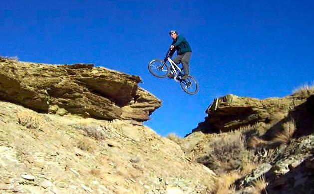 A mountain biker mid-air, leaping from one rocky cliff to another against a clear blue sky. The rugged terrain features sparse vegetation and steep rock formations, highlighting the thrill of the jump. Tnt mountain bike trail.
