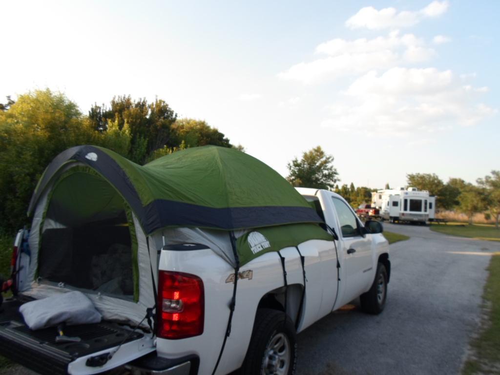 A white pickup truck with a green tent set up over the truck bed, parked on a gravel road surrounded by trees. In the background, additional vehicles and a camper can be seen, under a clear sky with scattered clouds. Alafia River State Park mountain bike trail.