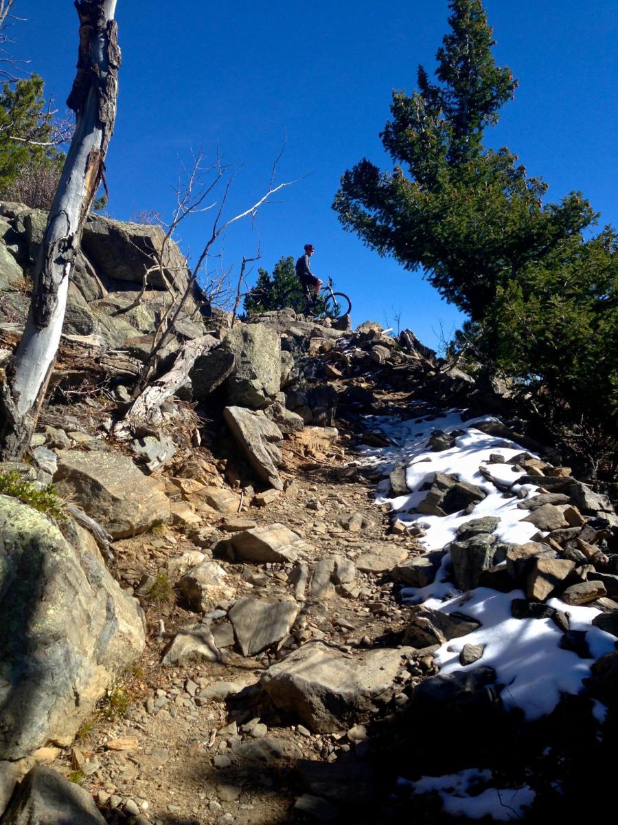 A mountain biker stands next to their bike on a rocky, steep trail surrounded by boulders and sparse vegetation, under a clear blue sky. Snow patches are visible along the path, suggesting a transition from winter to spring. Bergen Peak mountain bike trail.