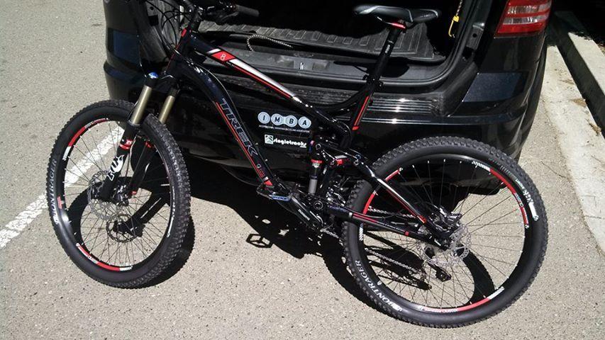Trek Fuel EX 8: A black and red mountain bike is parked next to an open trunk of a vehicle. The bike features thick tires, front suspension, and various stickers on the frame. The background shows a paved parking area under bright sunlight.