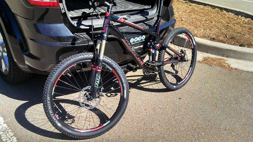 Trek Fuel EX 8: A mountain bike parked next to an open car trunk, featuring a black frame with red accents and distinctive wheels. The bike is positioned on a paved surface with a slight incline, and there is a patch of brown soil in the background.