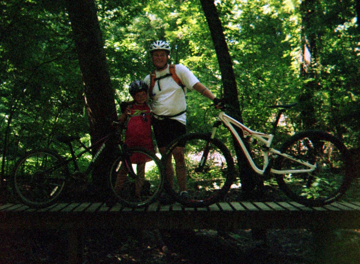 Specialized Camber Comp 29: A man and a child stand on a wooden bridge surrounded by dense greenery, each holding a mountain bike. The child wears a helmet and a vibrant outfit, while the man is dressed in athletic gear. They both smile, enjoying their outdoor biking adventure.