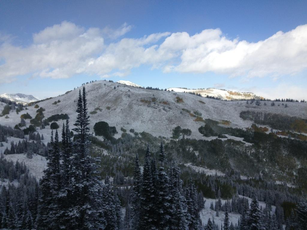 A snowy mountain landscape with rolling hills, tall evergreen trees in the foreground, and patches of autumn-colored foliage visible on the slopes. The sky is partly cloudy, featuring a mix of blue and white clouds. Dead Tree mountain bike trail.