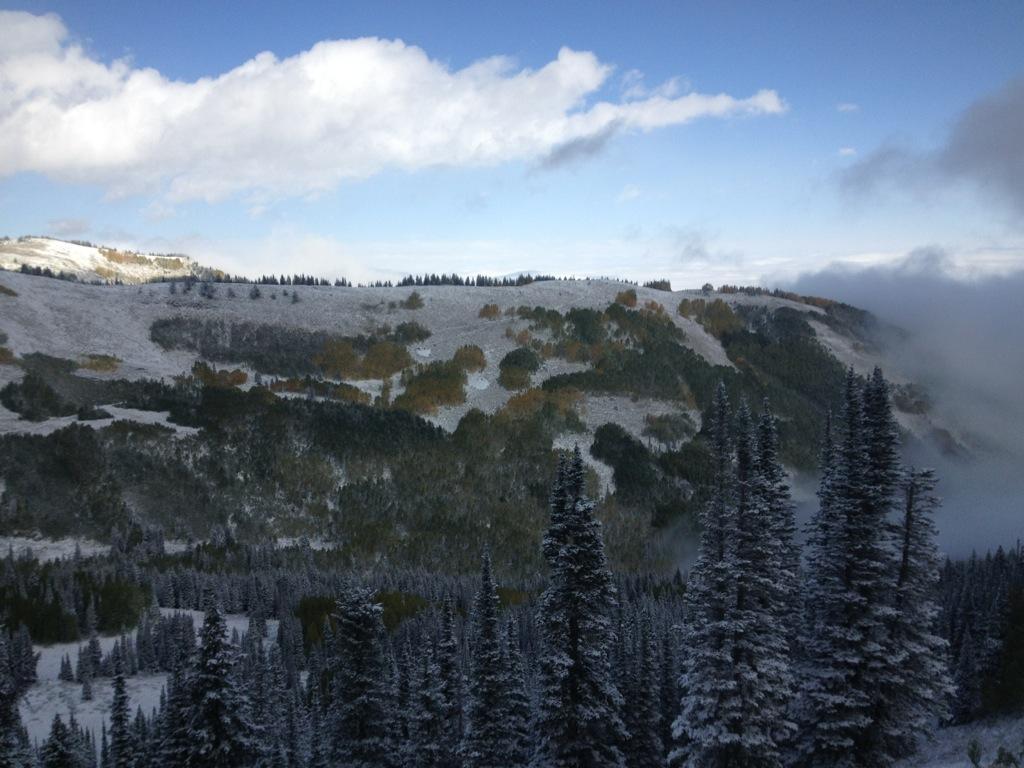 A panoramic view of a snow-covered mountainous landscape, featuring evergreen trees in the foreground and rolling hills in the background. The scene is brightened by a partly cloudy blue sky, with patches of snow on the ground and colorful autumn foliage visible on the hills. Dead Tree mountain bike trail.