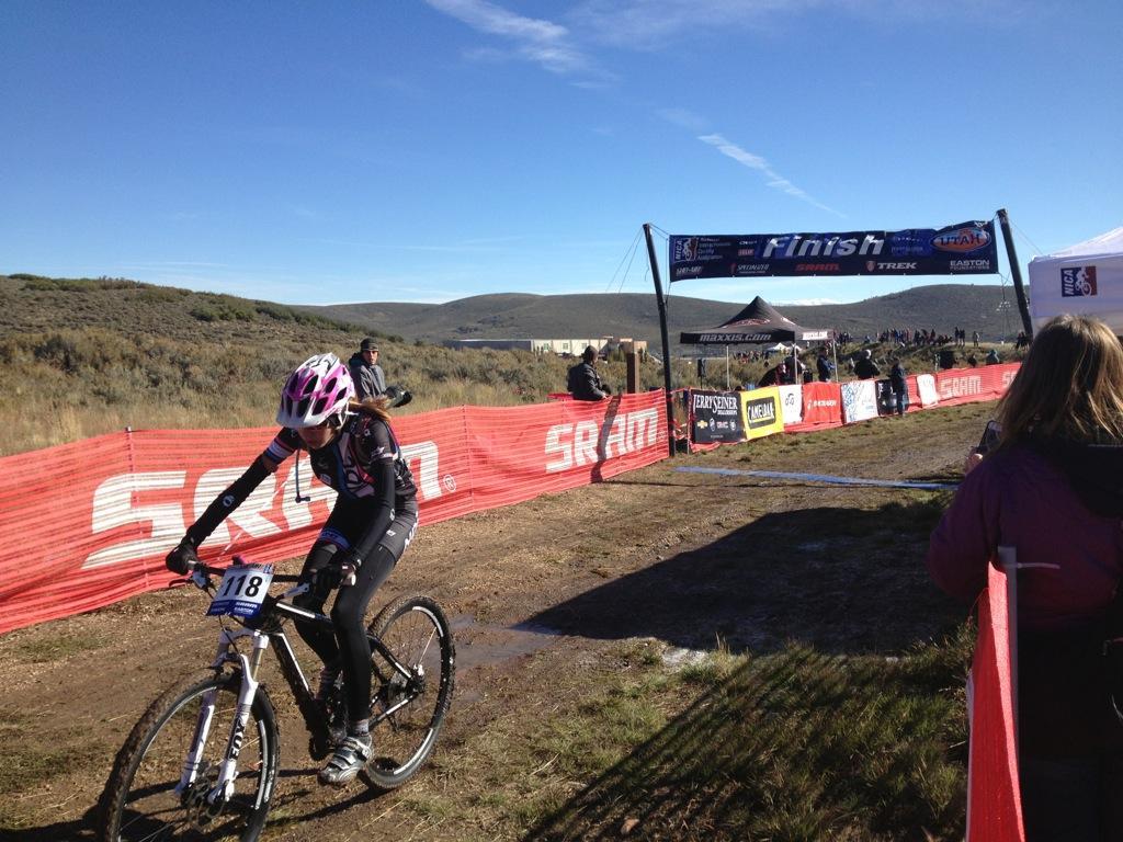 A cyclist in a black and pink outfit crosses the finish line of a mountain biking race, with a banner reading "Finish" overhead. The scene is set in an outdoor environment with spectators in the background and a blue sky. Red barriers surround the finish area. The cyclist is wearing a helmet and focused on completing the race. Round Valley mountain bike trail.