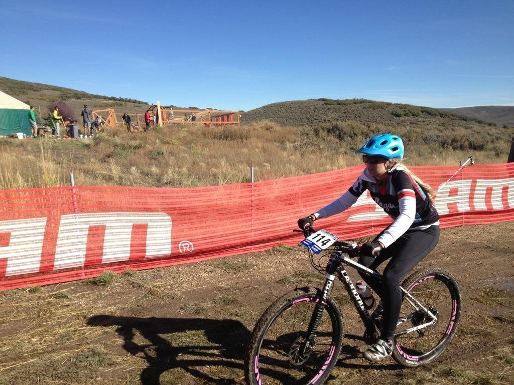 A young female mountain biker in a blue helmet and athletic gear rides her bike along a dirt path, with a red safety barrier in the foreground. In the background, a group of people is seen near a tent and wooden structure, surrounded by rolling hills and clear blue skies. The scene captures the energy of a cycling event. Round Valley mountain bike trail.