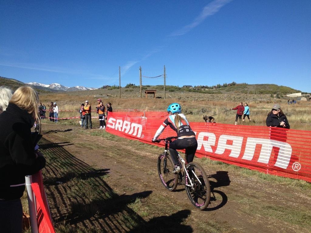 A female cyclist with a blue helmet is riding a mountain bike towards a finish line marked by red barriers displaying the SRAM logo. Spectators, including adults and children, are cheering from both sides of the track, with mountains visible in the background under a clear blue sky. Round Valley mountain bike trail.