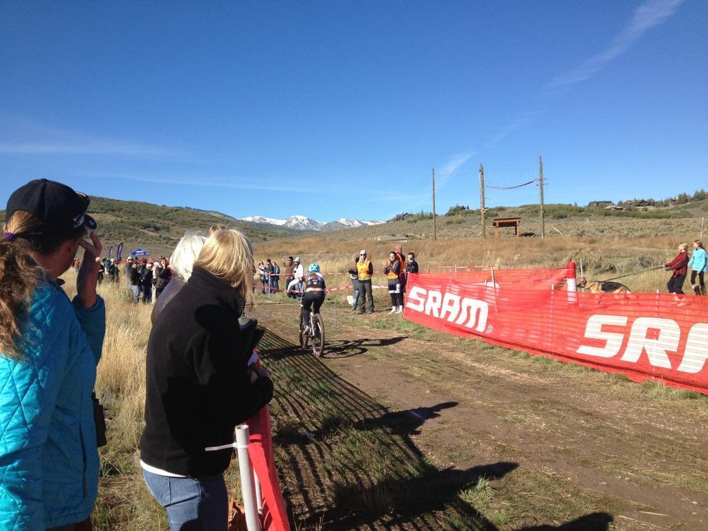 A mountain biker competes in a cycling race on a dirt trail, surrounded by spectators. The scene features a bright blue sky and distant snow-capped mountains. A red barrier advertising SRAM separates the biker from the crowd, with fans cheering and capturing the moment on their phones. Round Valley mountain bike trail.
