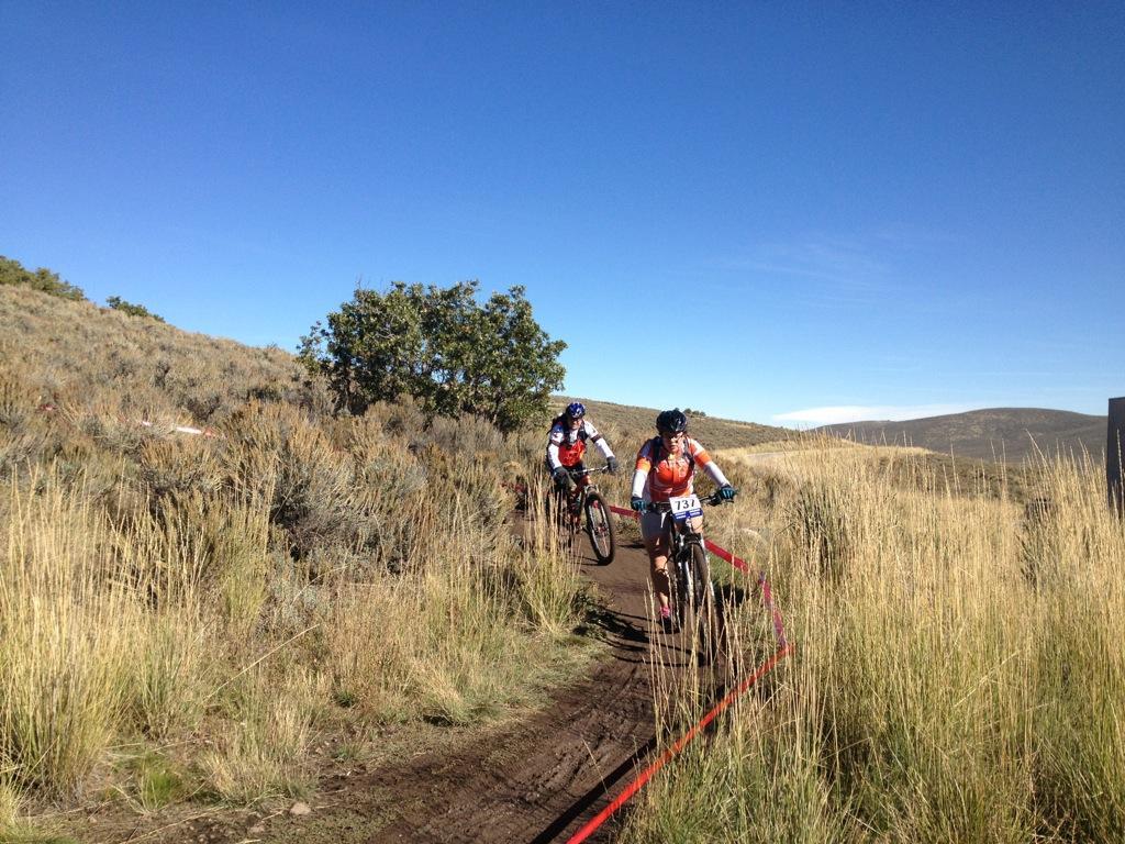 Two mountain bikers navigate a dirt trail surrounded by tall grass and shrubs. The scene is set under a clear blue sky, with rolling hills in the background. One biker is wearing a red jersey with a number 134, while the other is in a combination of red and black gear, showcasing their experience in a competition. A red ribbon marks the race path. Round Valley mountain bike trail.