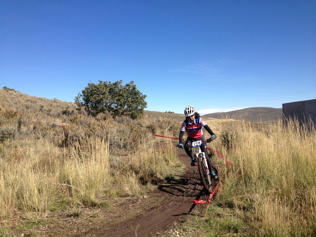 A mountain biker in a helmet and colorful jersey navigates a dirt trail, with tall grass and a small tree in the background under a clear blue sky. Red tape marks the course as they race through a competitive biking event. Round Valley mountain bike trail.