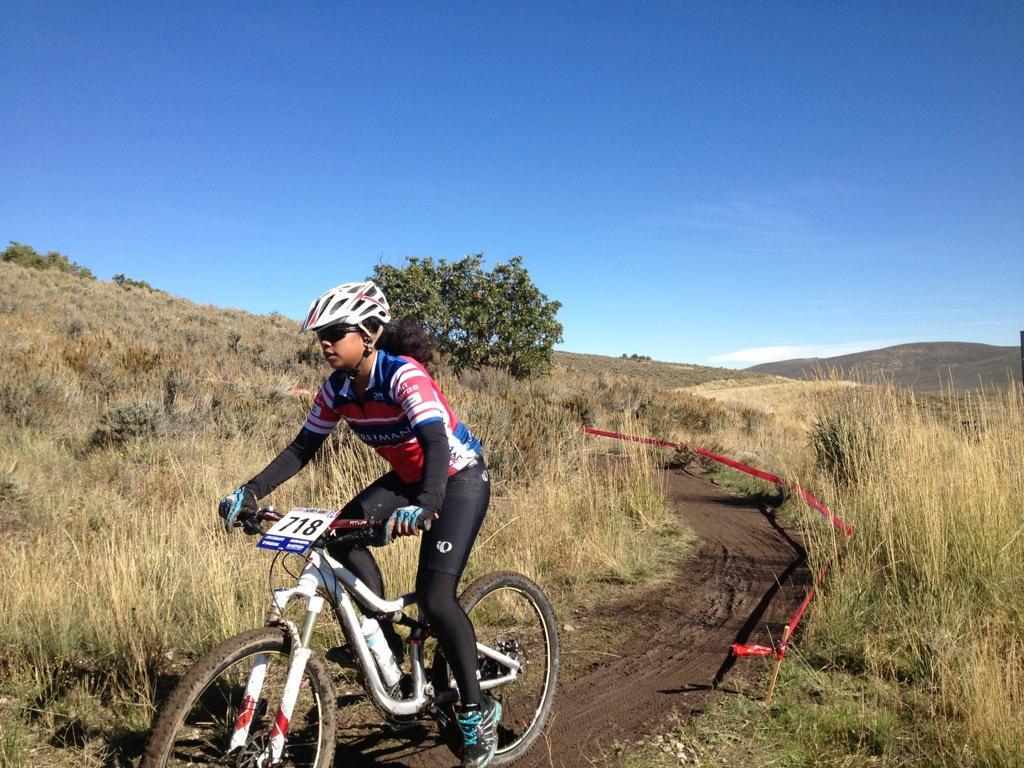A young female mountain biker with curly hair wears a helmet and cycling gear as she rides her bike along a dirt path in a grassy landscape. The trail curves to the right, marked by red tape, with rolling hills and a clear blue sky in the background. Round Valley mountain bike trail.