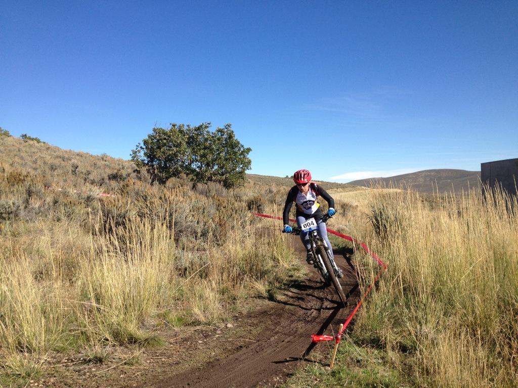 A mountain biker navigating a dirt trail surrounded by tall grass and shrubs, with a clear blue sky in the background. The cyclist is wearing a red helmet and racing gear, and is number 004. There are red course markers along the path. Round Valley mountain bike trail.