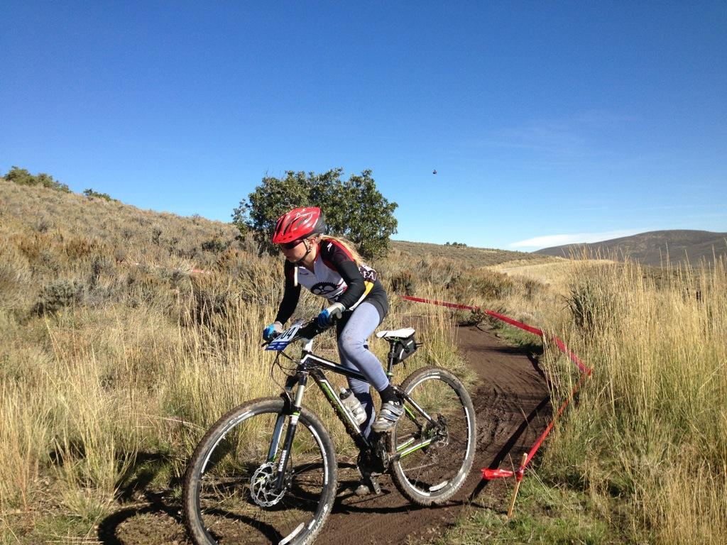 A cyclist riding a mountain bike on a dirt trail surrounded by tall grass and shrubs, under a clear blue sky. The cyclist is wearing a helmet and colorful attire, focused on navigating the trail while a red marker delineates the course. Round Valley mountain bike trail.