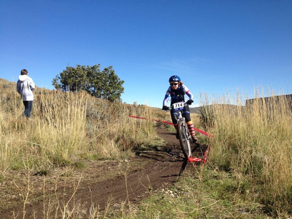 A cyclist wearing a blue helmet and racing jersey navigates a dirt trail, marked with red tape, through tall grass and shrubs. In the background, a person in a white sweatshirt watches the race under a clear blue sky. Round Valley mountain bike trail.