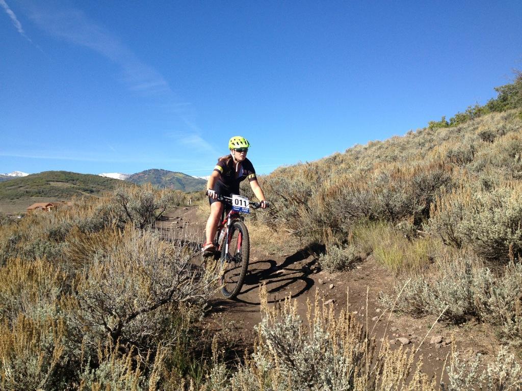 A cyclist wearing a helmet and race number 011 rides along a dirt trail surrounded by shrubs and grass, with mountains in the background under a clear blue sky. Round Valley mountain bike trail.