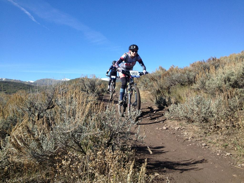 A mountain biker wearing a helmet and racing number 002 navigates a dirt trail surrounded by shrubs and hills under a clear blue sky. Another cyclist is seen in the background, contributing to the outdoor racing atmosphere. Round Valley mountain bike trail.