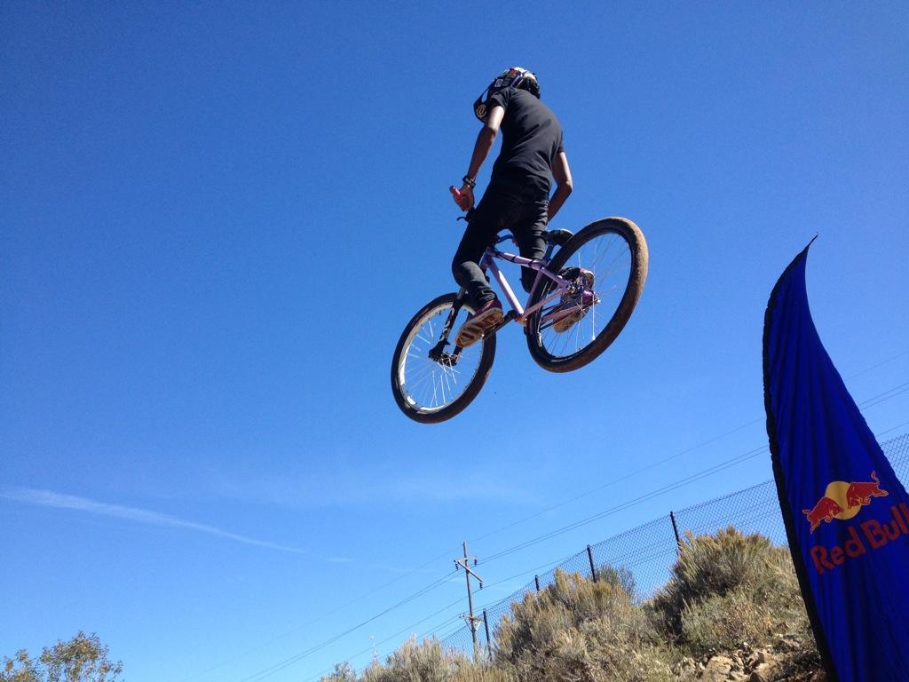A cyclist performing a jump on a bicycle in mid-air against a clear blue sky. The bike is pink, and the rider is wearing a black outfit and helmet. In the foreground, a Red Bull flag is visible, indicating a potential event or sponsorship. PC Dirt Jumps mountain bike trail.